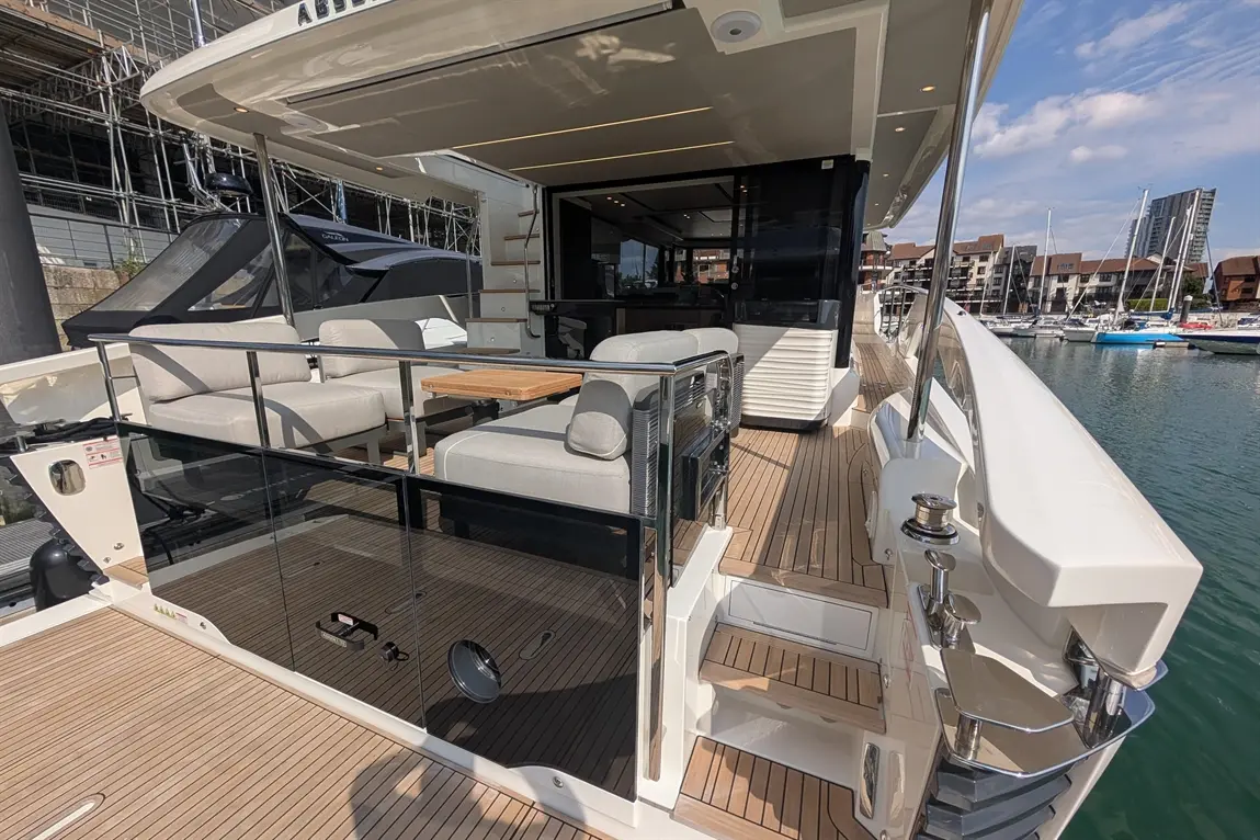 Luxury yacht deck with seating, wooden table, and glass railings, overlooking a marina with boats and nearby buildings.